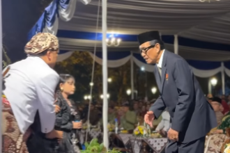 Man in a dark suit and traditional cap bows to a guest at a formal ceremony under a blue-and-white tent.