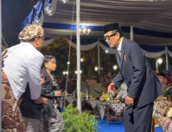 Man in a dark suit and traditional cap bows to a guest at a formal ceremony under a blue-and-white tent.