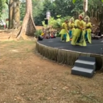 Outdoor cultural performance on a raised stage with dancers in yellow-green outfits, audience seated on brown couches to the left.