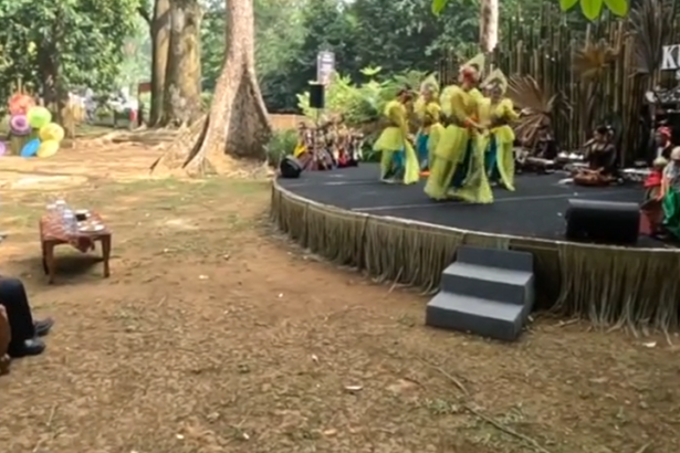 Outdoor cultural performance on a raised stage with dancers in yellow-green outfits, audience seated on brown couches to the left.