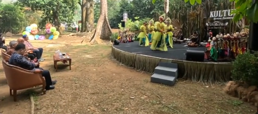 Outdoor cultural performance on a raised stage with dancers in yellow-green outfits, audience seated on brown couches to the left.