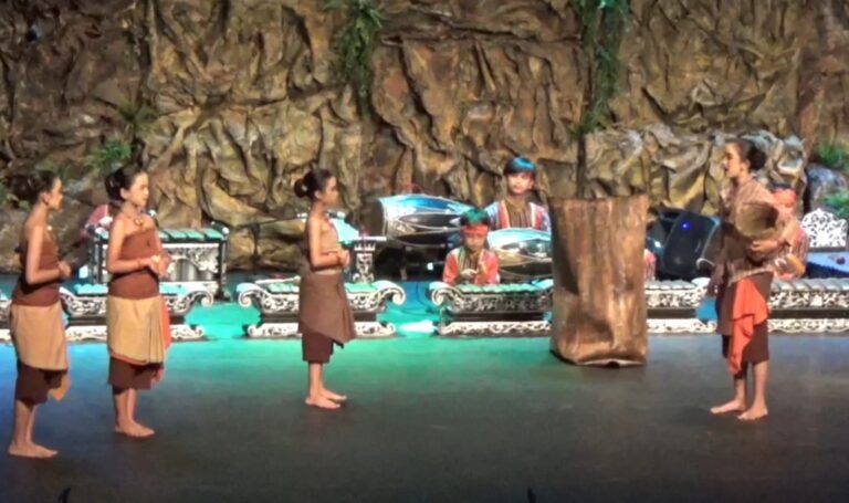 Group of young performers in brown costumes standing on a stage with a rocky backdrop, listening to a speaker on the right.