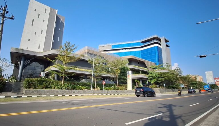 Modern multi-story building with blue glass facade along a busy street, with cars and motorcycles.