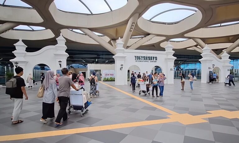 People waiting with luggage and carts outside Yogyakarta International Airport, white decorative arches and modern roof structure in the background.