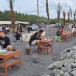 Groups of people sitting at small wooden tables on a dark sandy beach along a rocky breakwater, dining outdoors with drinks and snacks under a cloudy sky.