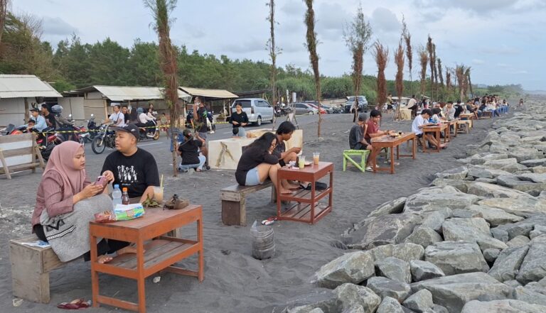 Groups of people sitting at small wooden tables on a dark sandy beach along a rocky breakwater, dining outdoors with drinks and snacks under a cloudy sky.