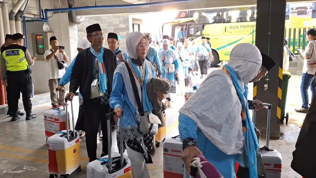 Group of Indonesian pilgrims in blue outfits and hijabs, pulling rolling suitcases through a terminal near a yellow tour bus labeled 'BUS PARIWIS'.