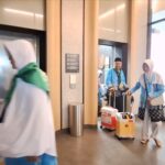 Two people wearing light-blue and white outerwear push wheeled suitcases through a modern lobby with elevator doors nearby.