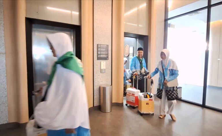 Two people wearing light-blue and white outerwear push wheeled suitcases through a modern lobby with elevator doors nearby.