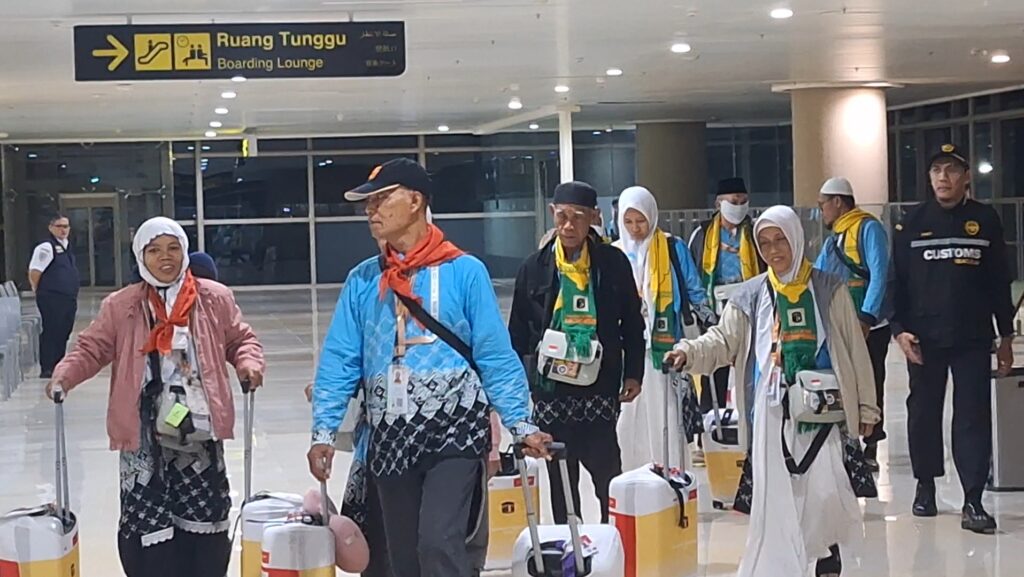 Group of travelers with rolling suitcases walking through an airport terminal, signposted for boarding lounge above them.