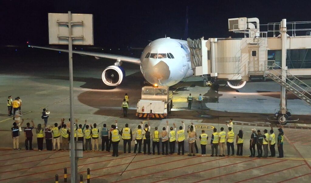 Airplane at gate at night with jet bridge attached and ground crew in yellow vests waving on the tarmac