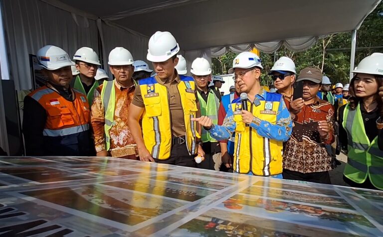 Group of construction workers and officials in hard hats and safety vests gathered around a large table map while a man with a microphone explains.