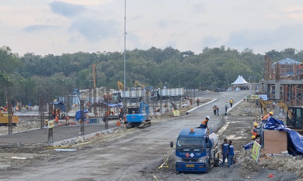 Construction site with workers, excavators, steel frames, and trucks against a forest backdrop