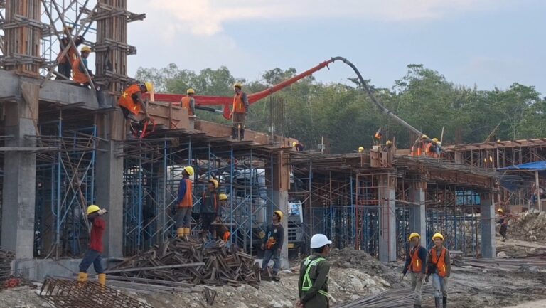 Construction workers in yellow hard hats and orange vests stand on scaffolding as a concrete pump pours into a building frame.