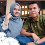 Smiling couple posing for a selfie indoors: woman wearing a blue hijab and patterned white dress, man in a green polo shirt beside her.