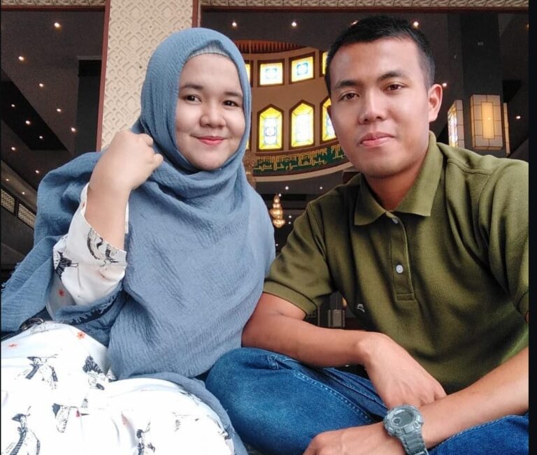 Smiling couple posing for a selfie indoors: woman wearing a blue hijab and patterned white dress, man in a green polo shirt beside her.