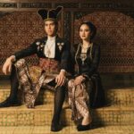A man and woman in traditional Indonesian ceremonial dress sit together on tiled steps with ornate batik fabrics and jewelry behind them.