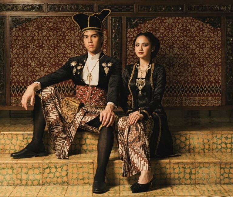 A man and woman in traditional Indonesian ceremonial dress sit together on tiled steps with ornate batik fabrics and jewelry behind them.