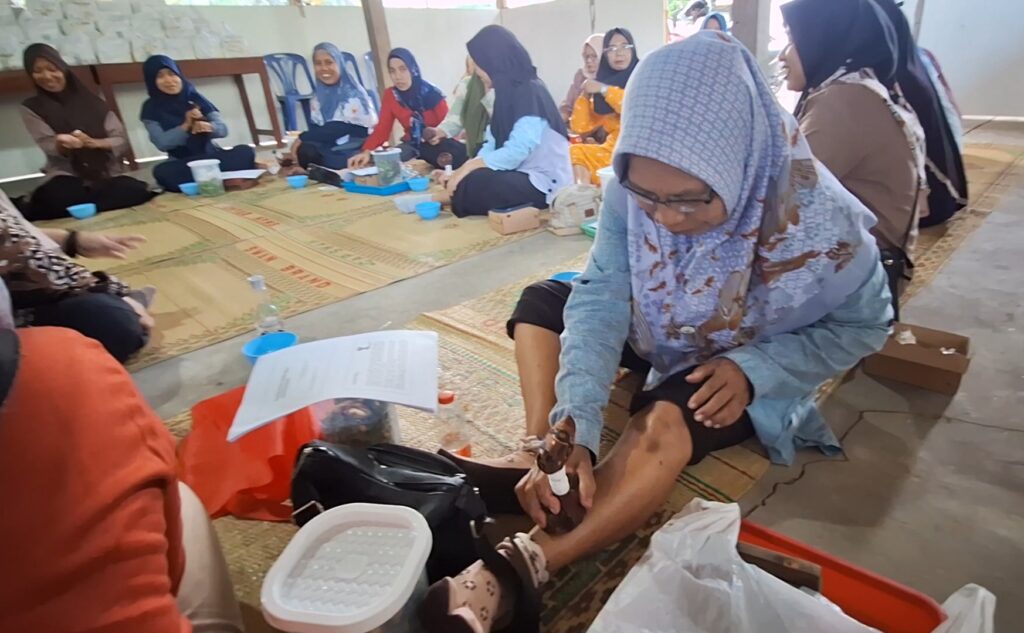 Group of women in hijabs seated on a mat in a circle, participating in a foot care workshop; a woman at the front applies product to a participant's foot.
