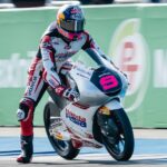 Moto racer in Red Bull helmet leaning into a turn on a Honda Asia race bike with white-red livery, trackside green barrier in background