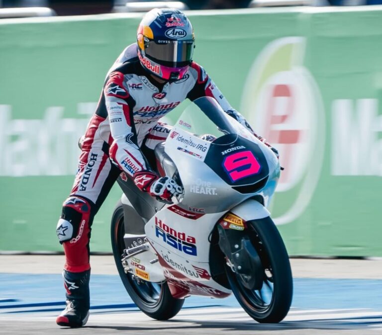 Moto racer in Red Bull helmet leaning into a turn on a Honda Asia race bike with white-red livery, trackside green barrier in background