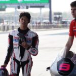MotoGP rider in a white Honda racing suit stands with helmet in hand beside a crew member at the pit lane.