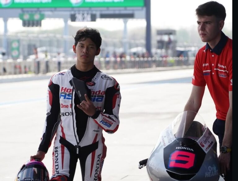 MotoGP rider in a white Honda racing suit stands with helmet in hand beside a crew member at the pit lane.