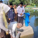 Group of officials and a bride-like guest perform a ritual by a pond; a man in white bows toward the water while others watch.