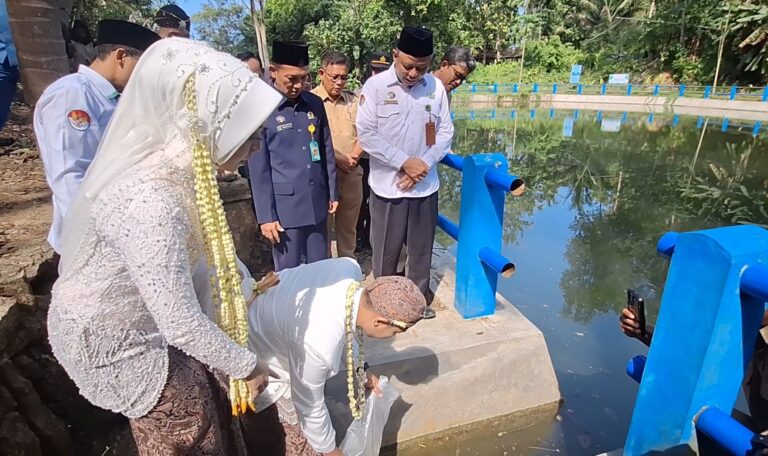 Group of officials and a bride-like guest perform a ritual by a pond; a man in white bows toward the water while others watch.