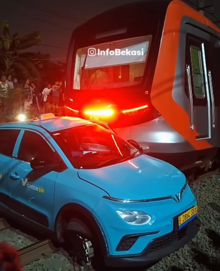 Blue SUV with a 'GREEN SM' logo sits on tracks next to a stationary orange-and-gray train at night, red emergency lights flashing and onlookers nearby.