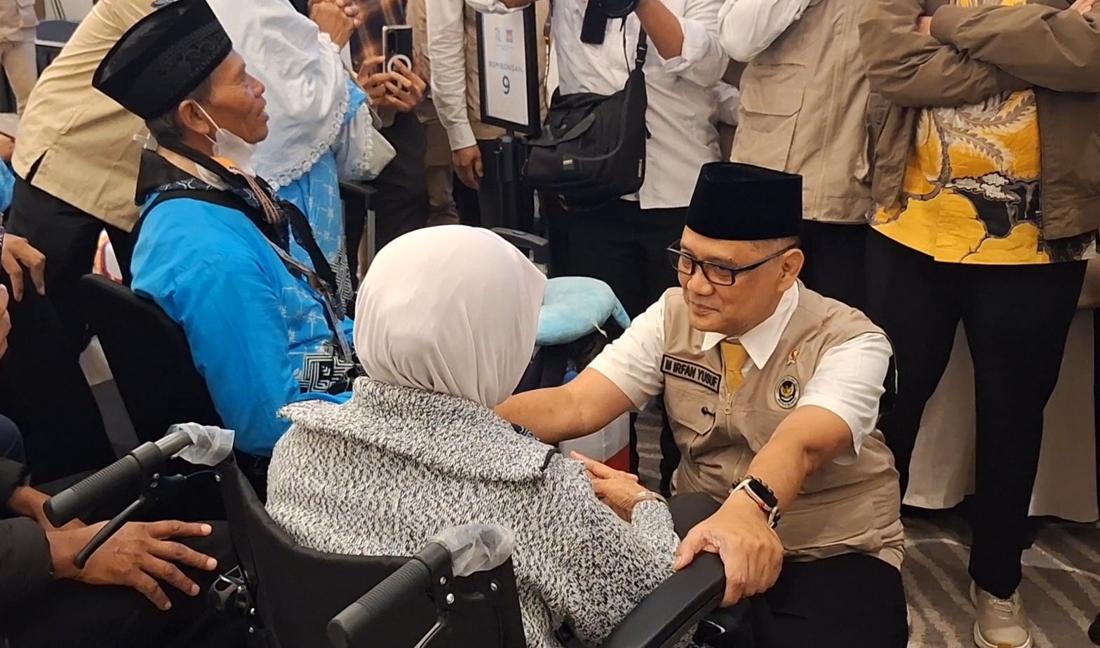 A man in a beige uniform and black cap kneels beside an elderly woman in a wheelchair, holding her hands in a crowded room. The man’s name tag reads ‘Irfan Yusof.’