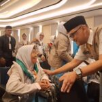 Volunteer in a tan uniform greets an elderly woman in a wheelchair at a formal event hall.