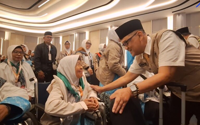 Volunteer in a tan uniform greets an elderly woman in a wheelchair at a formal event hall.