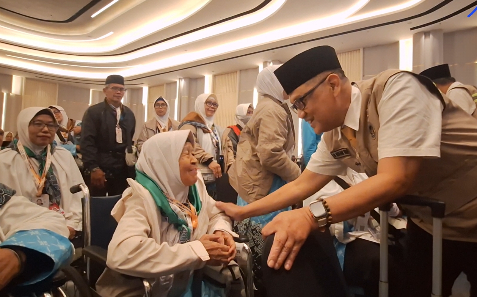 Volunteer in a tan uniform greets an elderly woman in a wheelchair at a formal event hall.