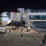 White commercial airplane docked at a jet bridge at night, ground crew in reflective vests nearby.