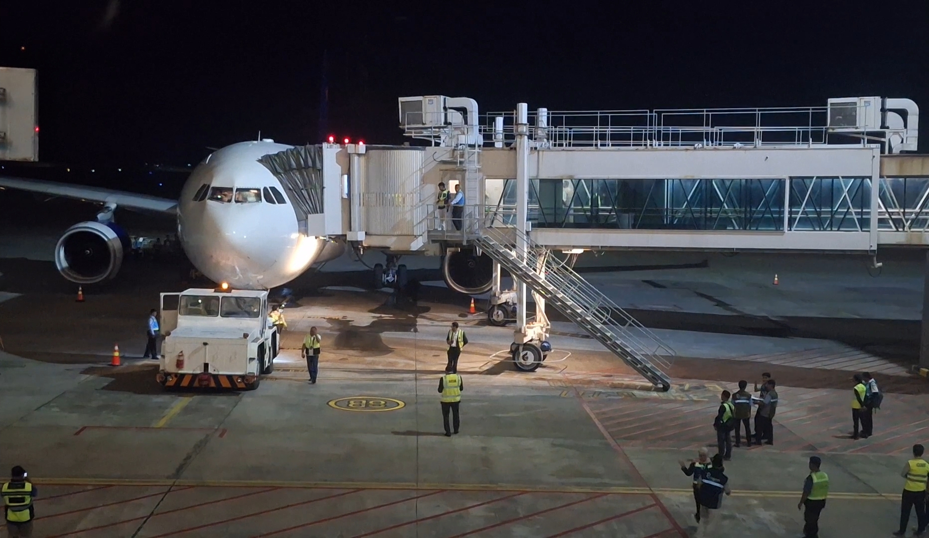 White commercial airplane docked at a jet bridge at night, ground crew in reflective vests nearby.