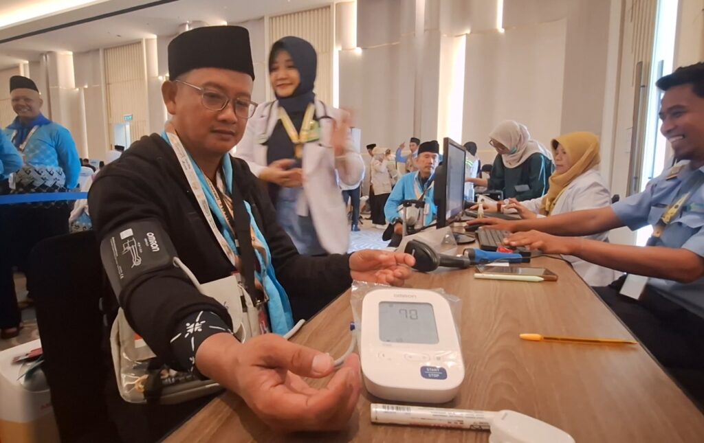 Man in a black hat has his blood pressure checked with an Omron cuff at a health screening table, staff and attendees in background.