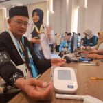 Man in a black hat has his blood pressure checked with an Omron cuff at a health screening table, staff and attendees in background.