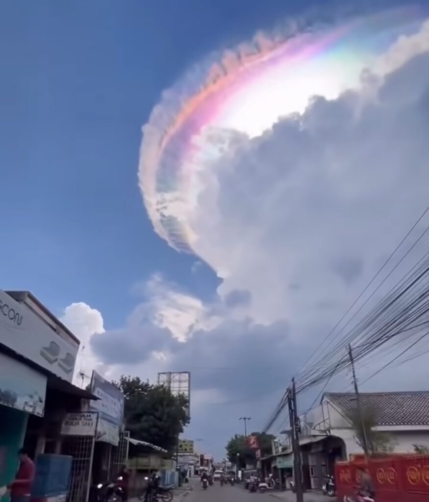Ring-shaped iridescent cloud arc forming a rainbow circle in a blue sky above a street and buildings.