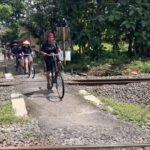 Group of cyclists crossing a rail crossing on a rural path, leading rider wearing red headscarf and black shirt, others follow behind.