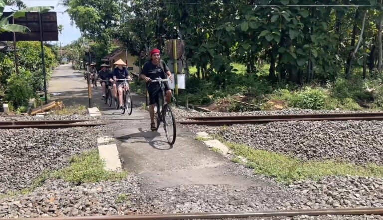 Group of cyclists crossing a rail crossing on a rural path, leading rider wearing red headscarf and black shirt, others follow behind.