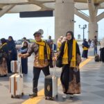 Group of travelers with yellow scarves walking through a modern airport terminal with luggage in tow under a concrete canopy.