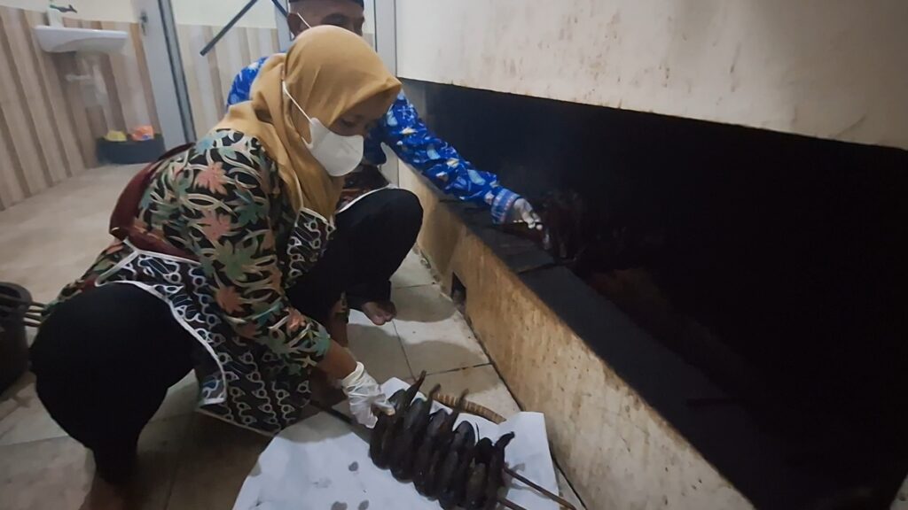 Two women wearing masks and gloves crouch by a counter, cleaning a large fish laid on a white cloth in a kitchen area.