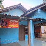 Bright blue house with a brown door and a red banner reading 'KAMPUNG LELE ASAP' hanging above the entrance.
