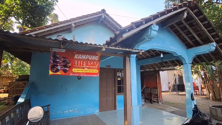 Bright blue house with a brown door and a red banner reading 'KAMPUNG LELE ASAP' hanging above the entrance.