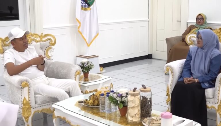 Two people sit on ornate white-and-gold couches across a small table in a bright room, casually talking; man in white cap and T-shirt on the left, two women in hijabs on the right.