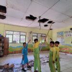 Four students in green and yellow uniforms stand in a classroom, holding a long wooden pole to reach a damaged ceiling with several holes.