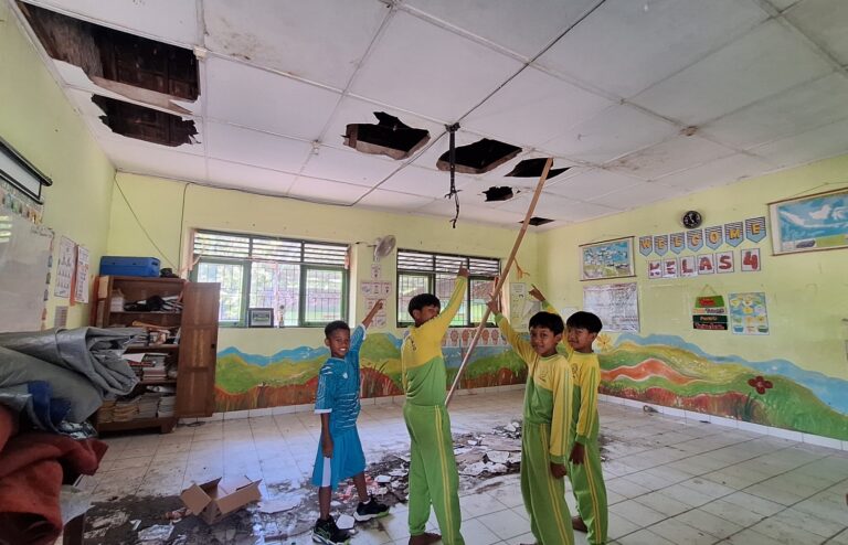 Four students in green and yellow uniforms stand in a classroom, holding a long wooden pole to reach a damaged ceiling with several holes.