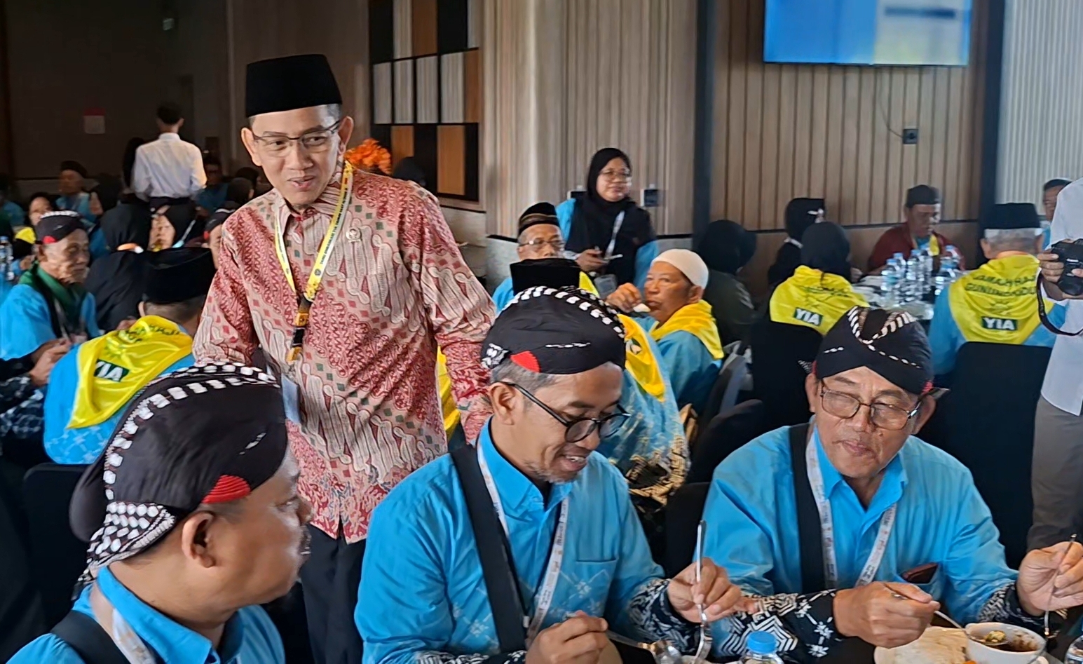 Group of men in blue shirts with lanyards and traditional headwear dining at a conference banquet in a crowded hall.