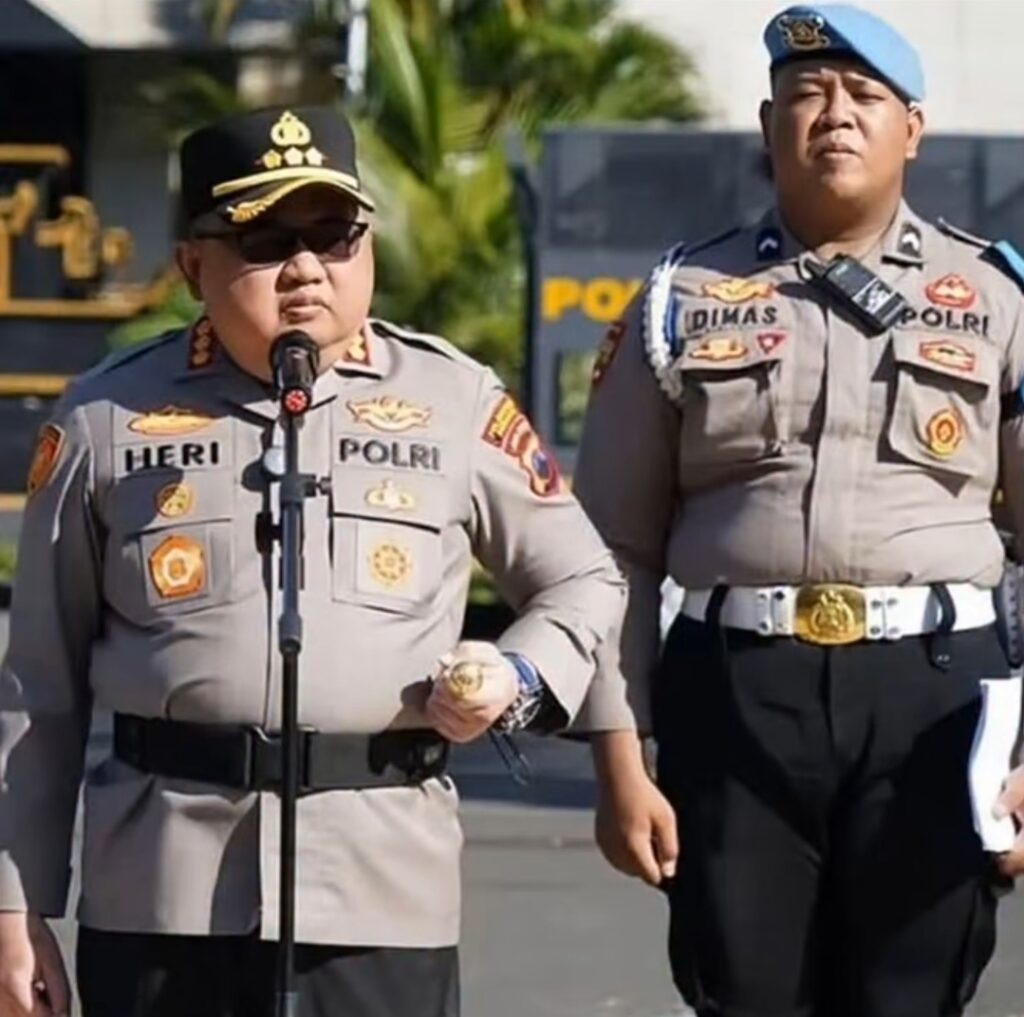 Two Indonesian police officers in khaki uniforms stand outdoors; one speaks into a microphone while the other looks on.